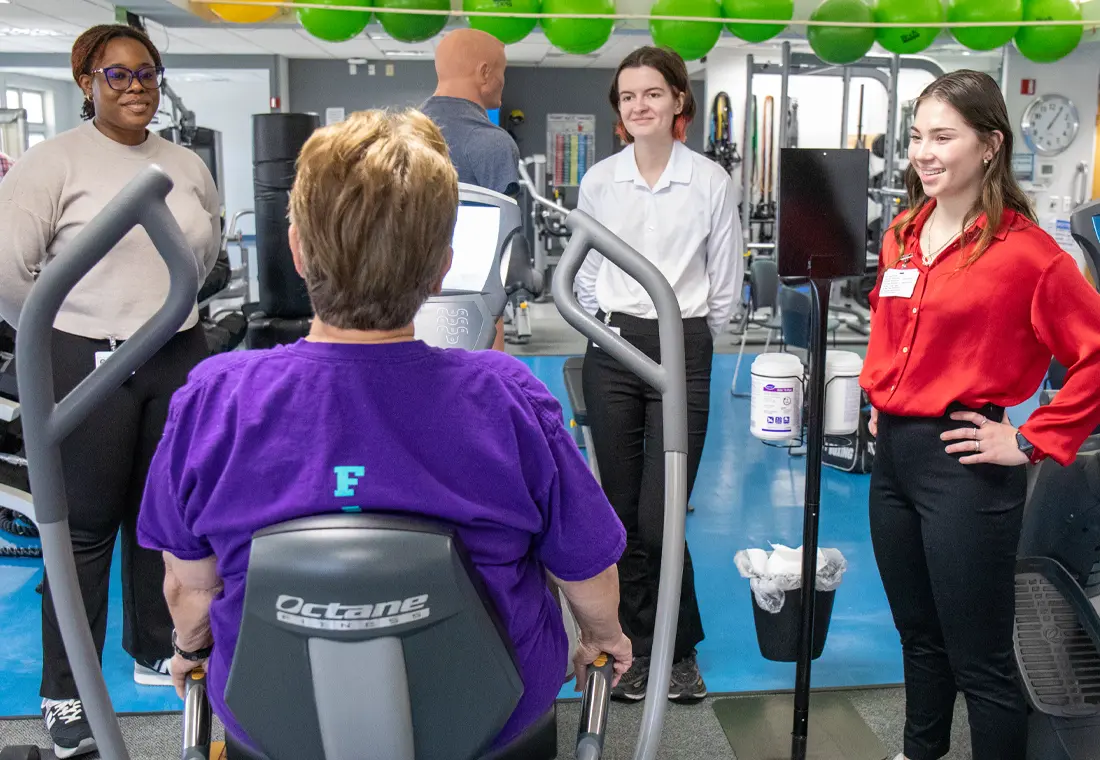 In a gym environment, three individuals stand and smile at a woman using fitness equipment.