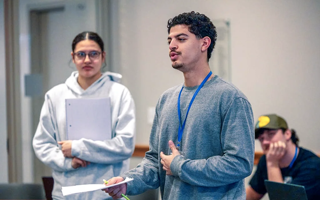 A student stands in front of a classroom, speaking. Two students are visible in the background.
