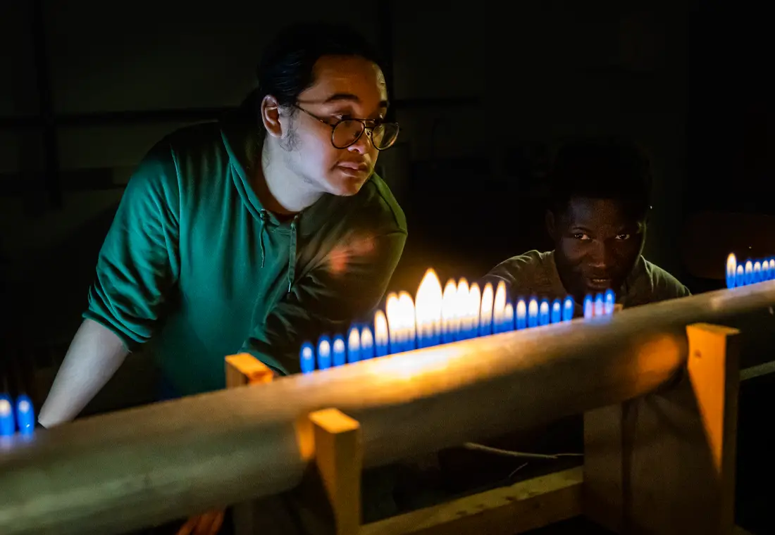 Two students observe multiple flames, some of which are taller and bigger than others. All the flames have a blue hue at the bottom, turning toward yellow at the tops.