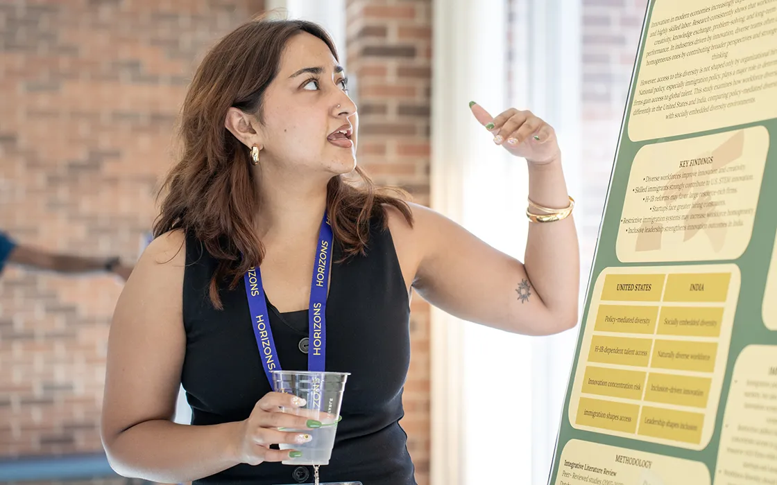 A student wearing a Horizons lanyard points at her presentation poster as she explains the concept of her research.