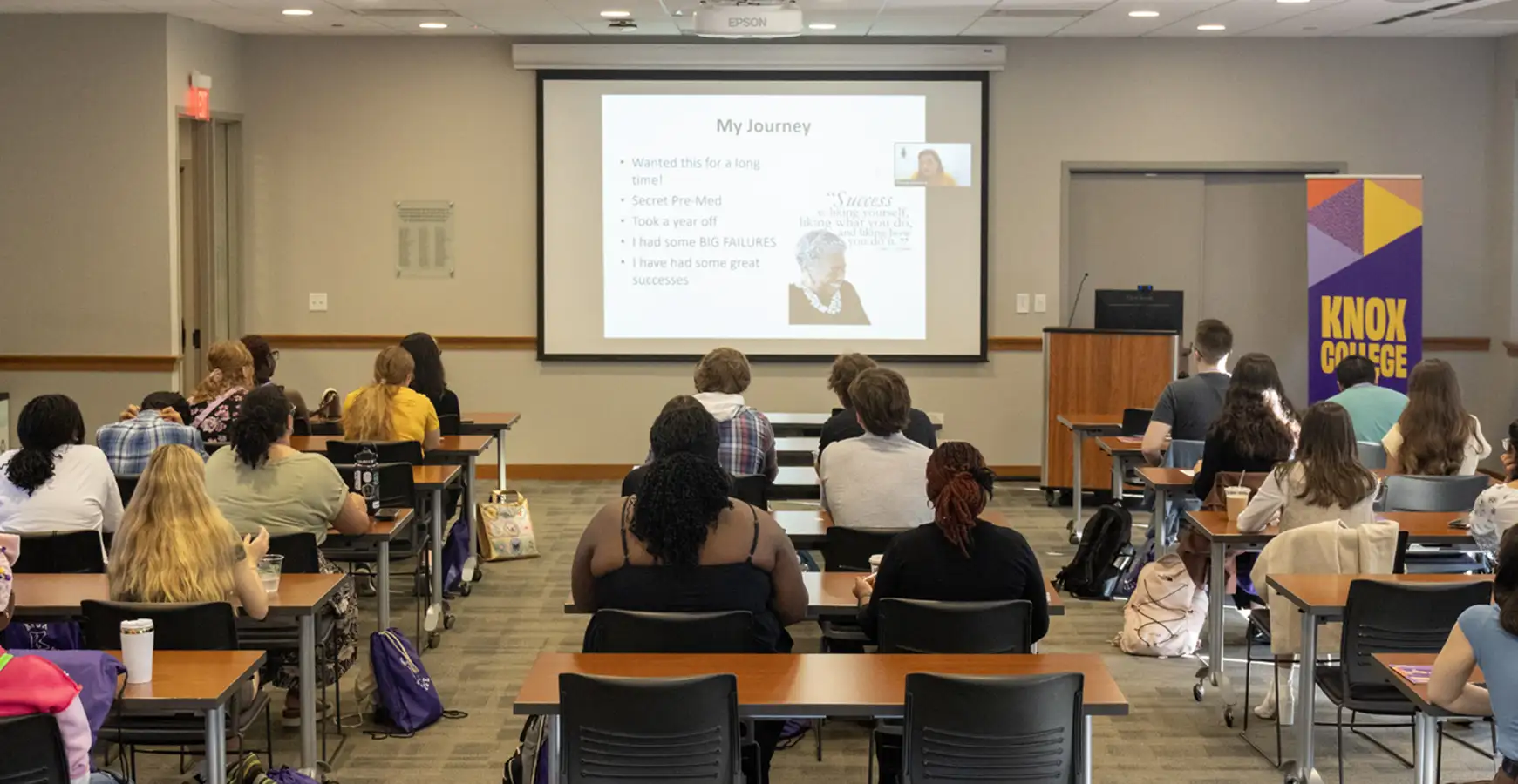 Students sit at tables appearing to be taking notes while a speaker presents.