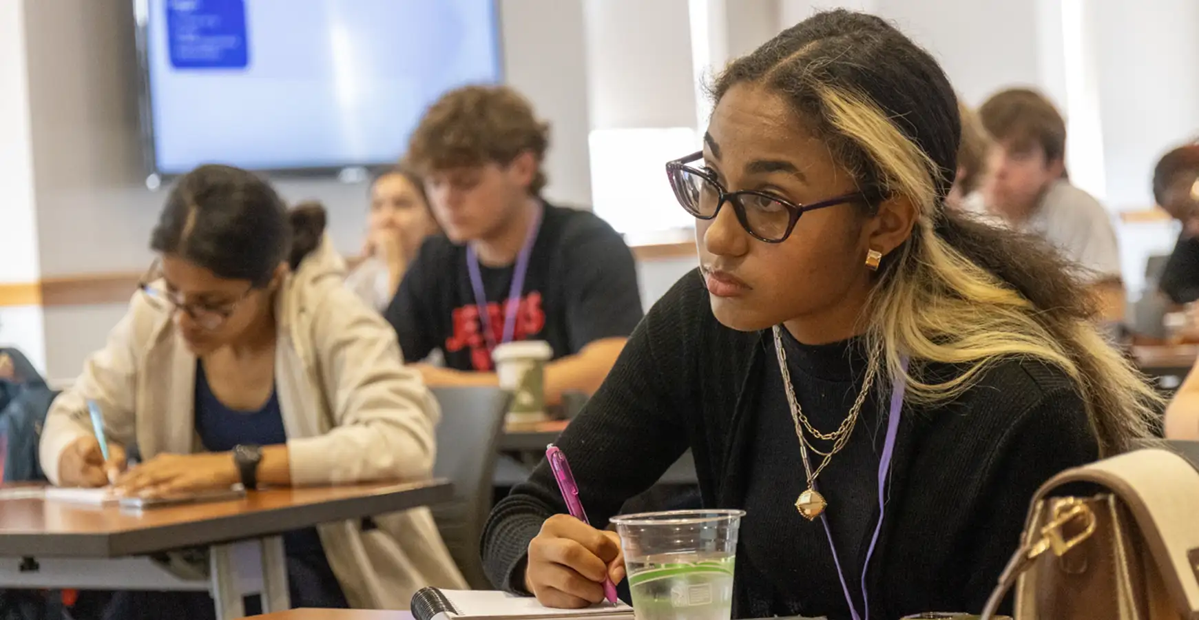 Students sit at tables appearing to be taking notes while a speaker presents.