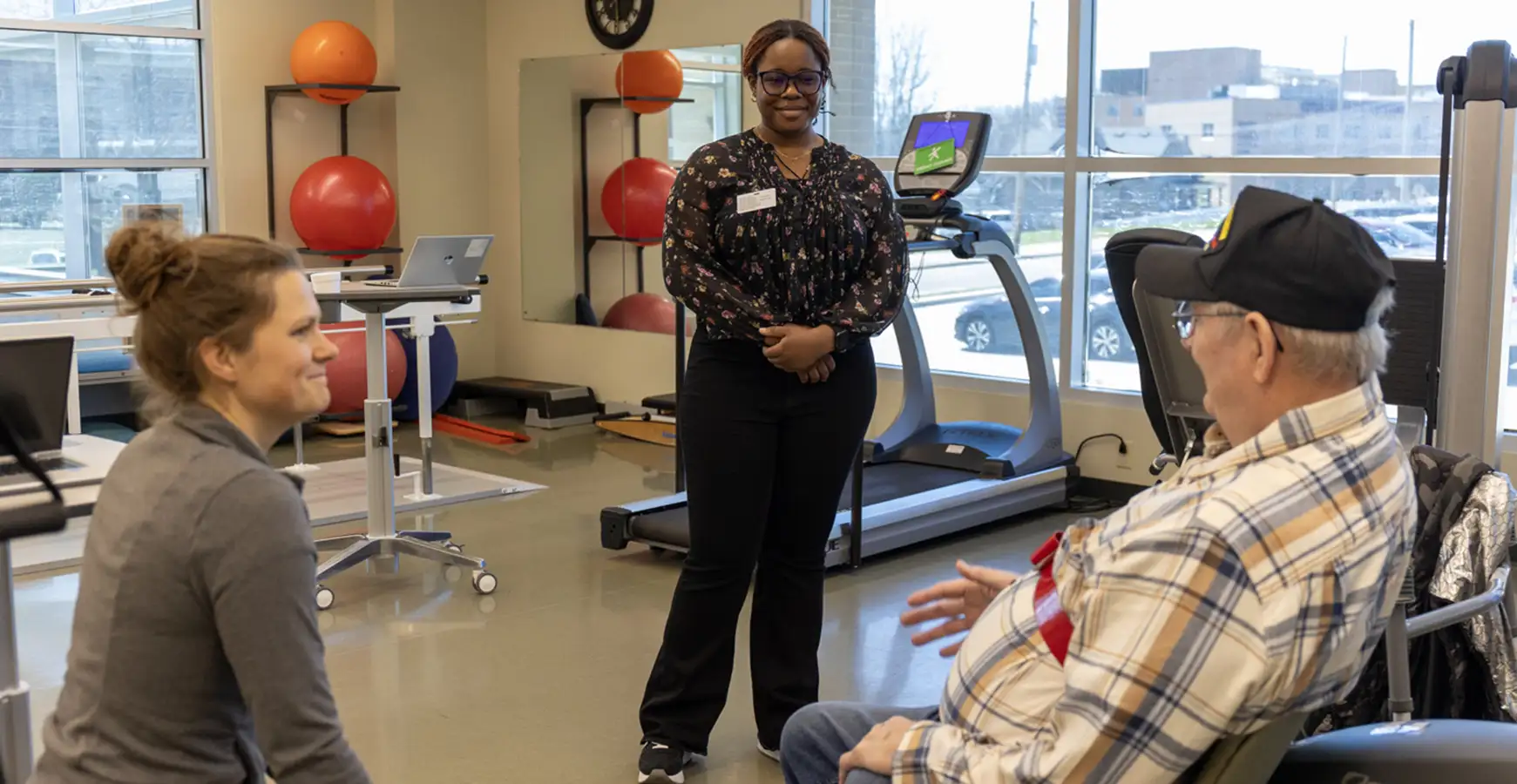 A student stands in front of an employee she is shadowing and a patient who is sitting down in a wheelchair.