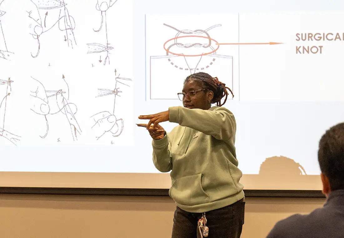 A student stands in front of a white board with a presentation of surgical knots in the background.