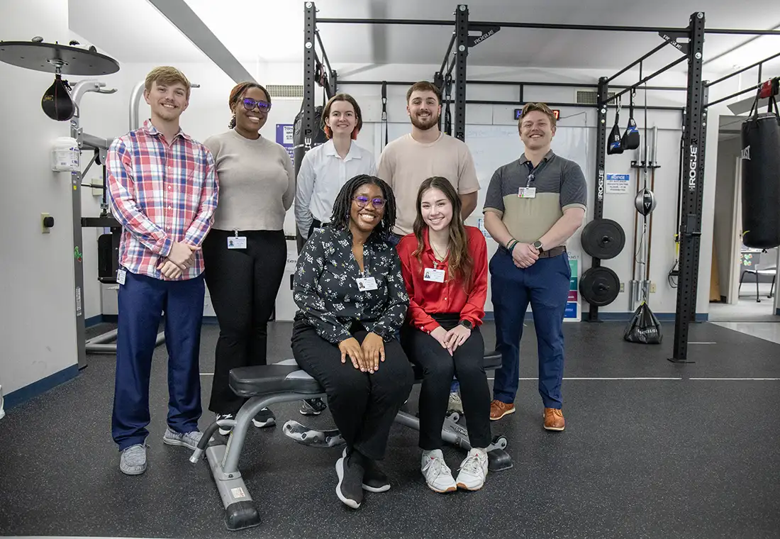 A group of students sit in a physical therapy department and smile at the camera.
