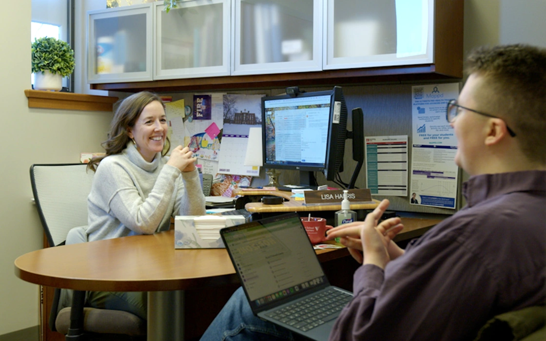 Lisa Harris smiles as a person sits across from her with a laptop open and speaks.