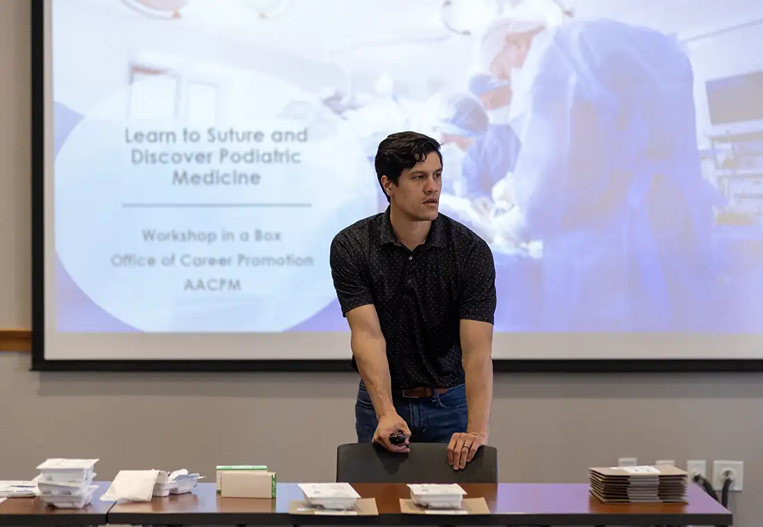 An instructor stands in front of a room with a presentation on suturing on a screen behind him.