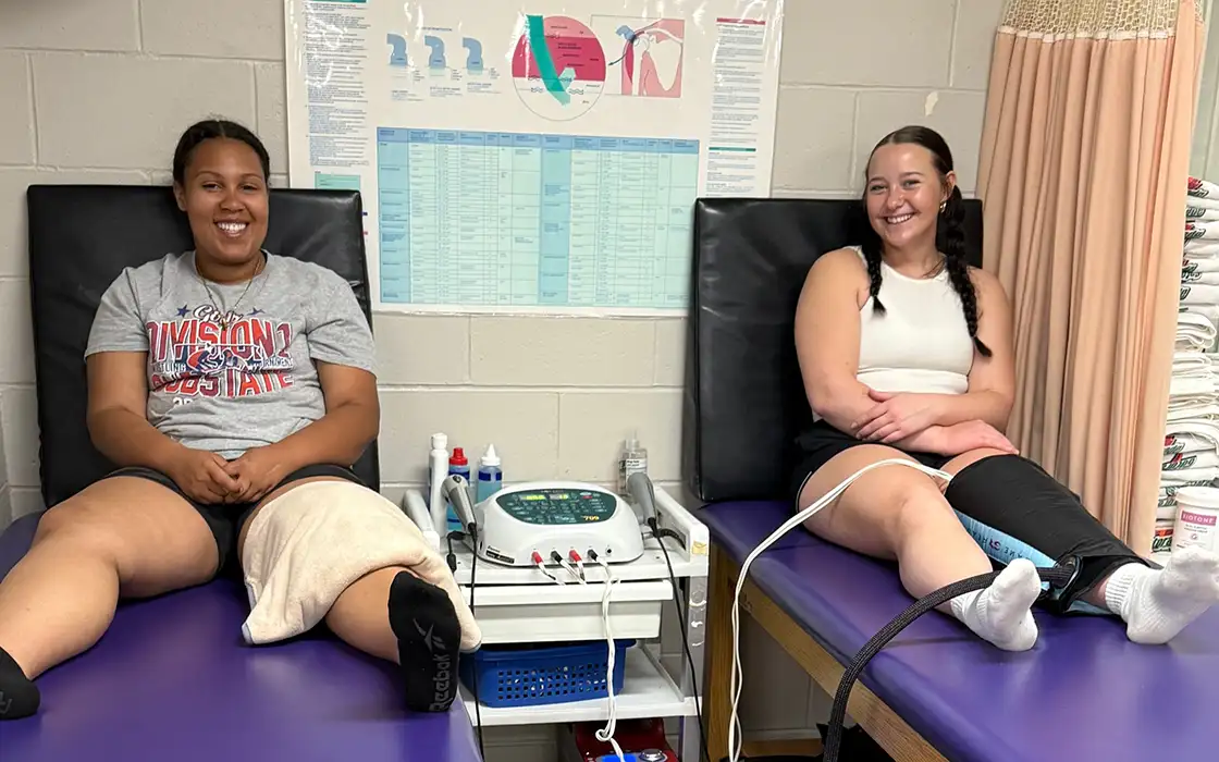 Two students each sit on an exam table with wraps on their legs.