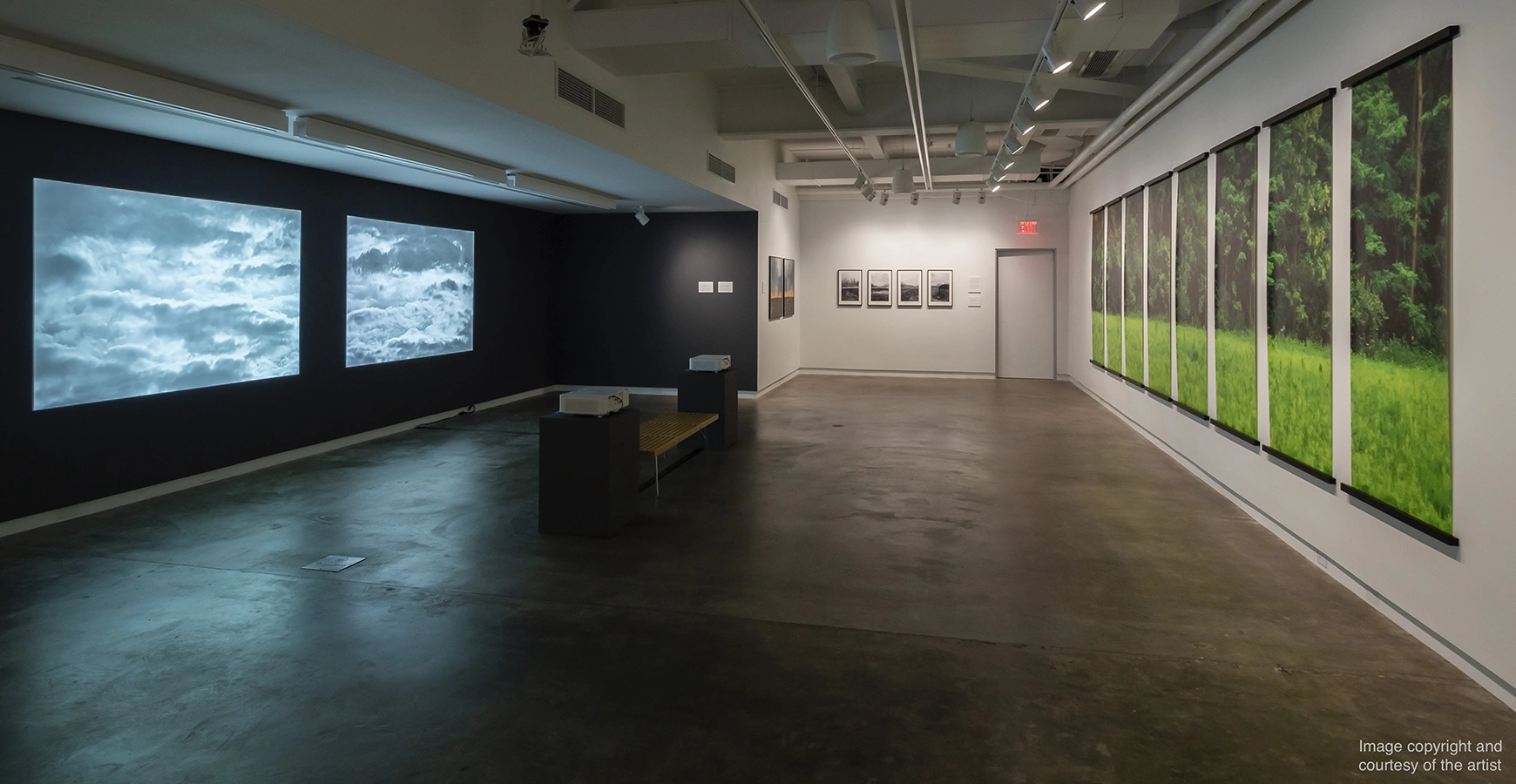 A wide open room is dimly lit. Two screens showing clouds are on the wall on the left, and images of a prairie are visible on the right. Text on the image says Image copyright and courtesy of the artist. 