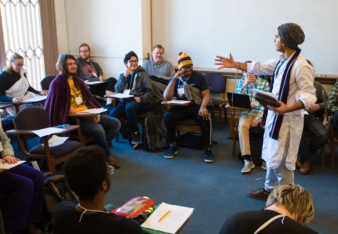 Students in a classroom smile at a speaker, who holds a book in his hand, a rosary dangles from his wrist, he wears a kurta, a shawl, and a knit cap.