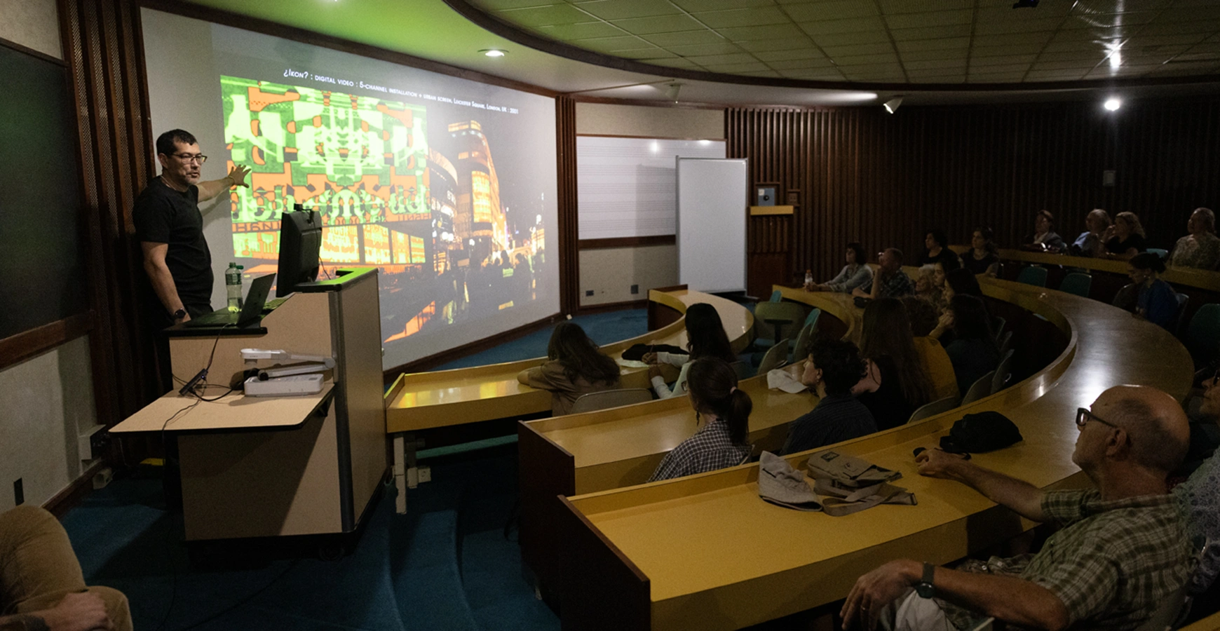 A man presents images of art on a screen. An audience is visible in front of him. 