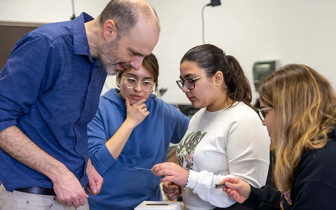A professor smiles as he leans in to examine what a student presents to him; two other students look on.