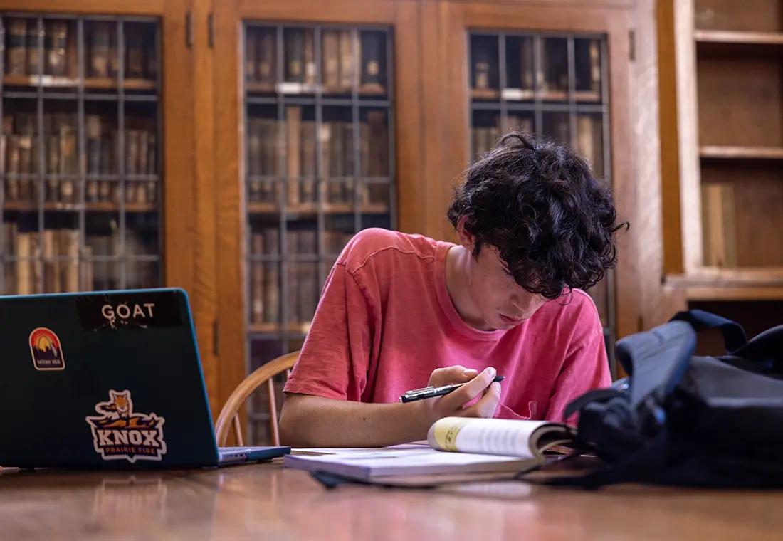 A student studies in Seymour Library with a book and laptop spread on the table.