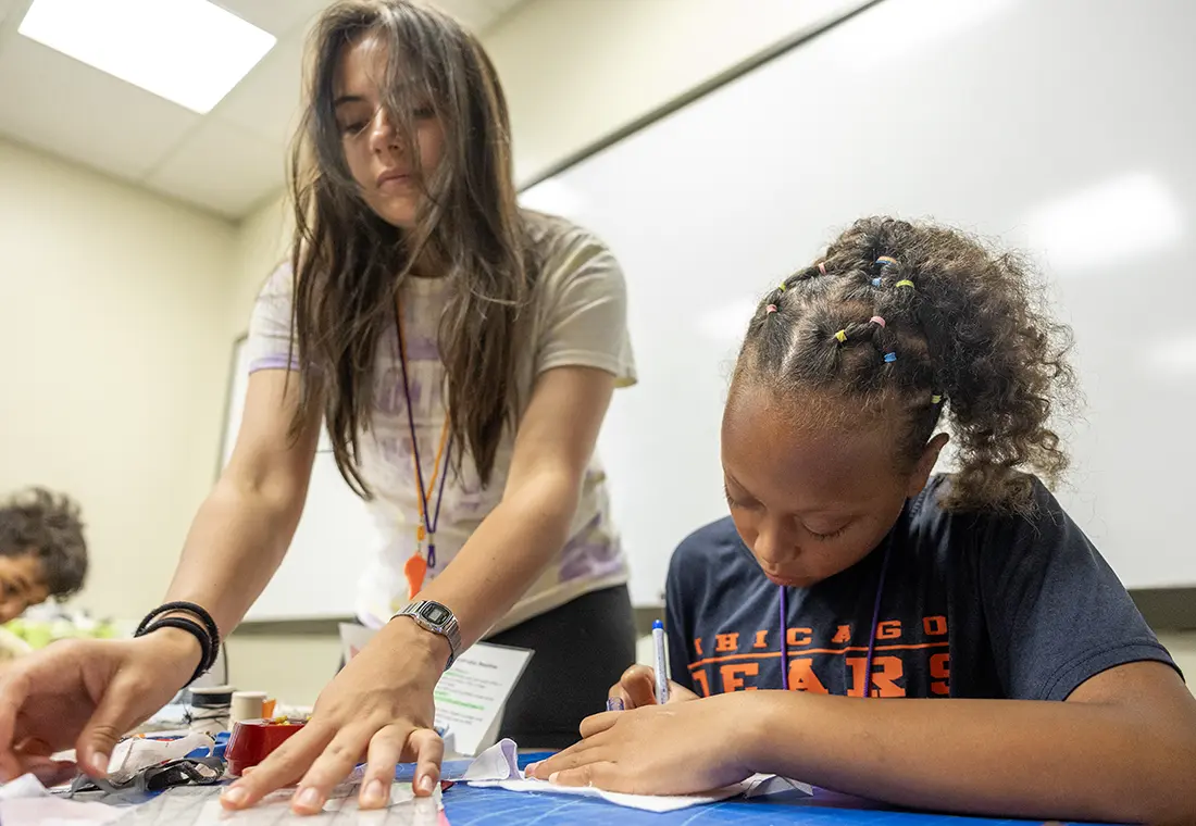 A woman wearing an orange whistle leans down to help a child with a project.