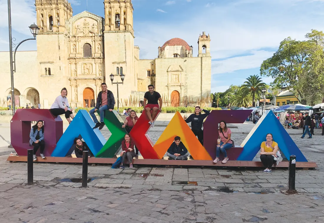 A group of students sit on large letters that spell out Oaxaca. Behind them, historic site Templo de Santo Domingo de Guzmán is visible. 