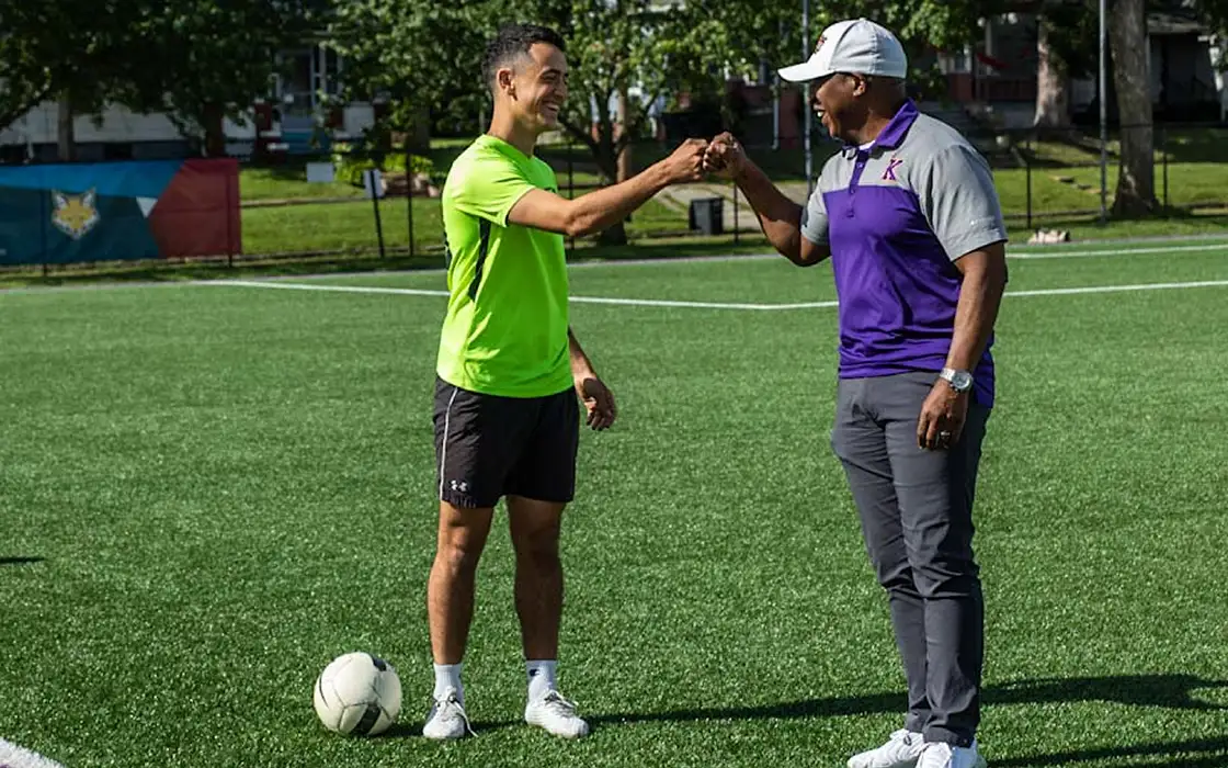 A student and a coach fist bump on the field. A soccer ball is on the ground near the student's foot.