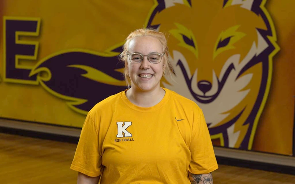 Coach Casey Hipp wears a yellow Knox softball shirt and smiles at the camera.