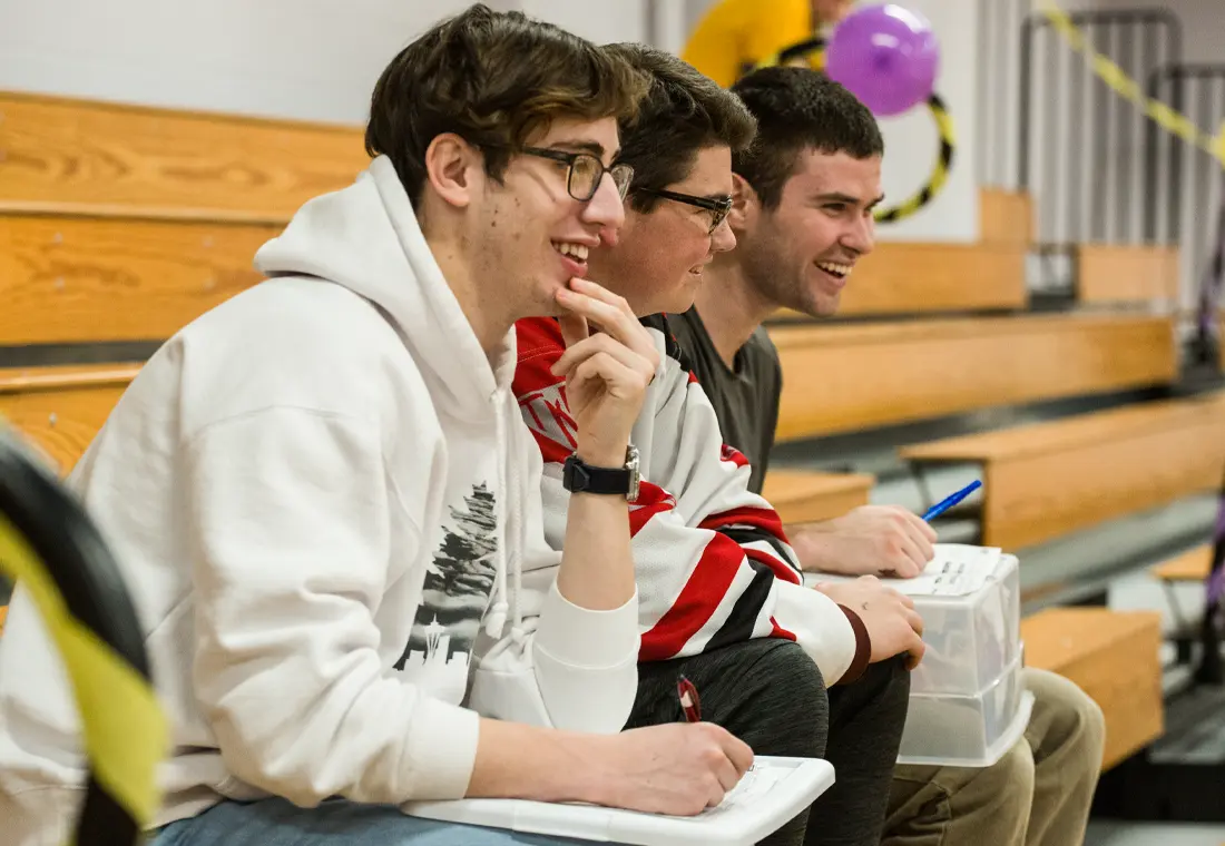 Three students sit on bleachers in a gymnasium setting. They all smile; two of them have paper and pen on their laps.
