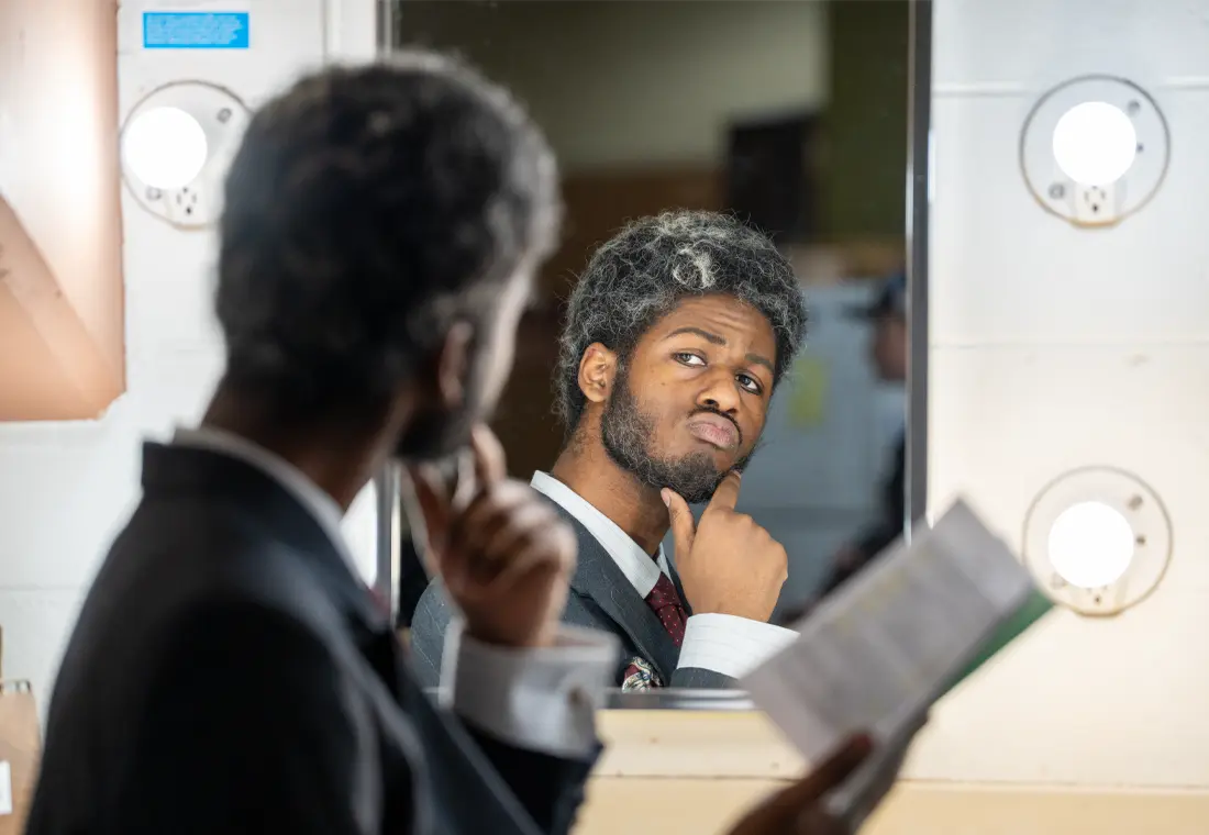 A student actor regards himself in the mirror, script in hand, before a performance.