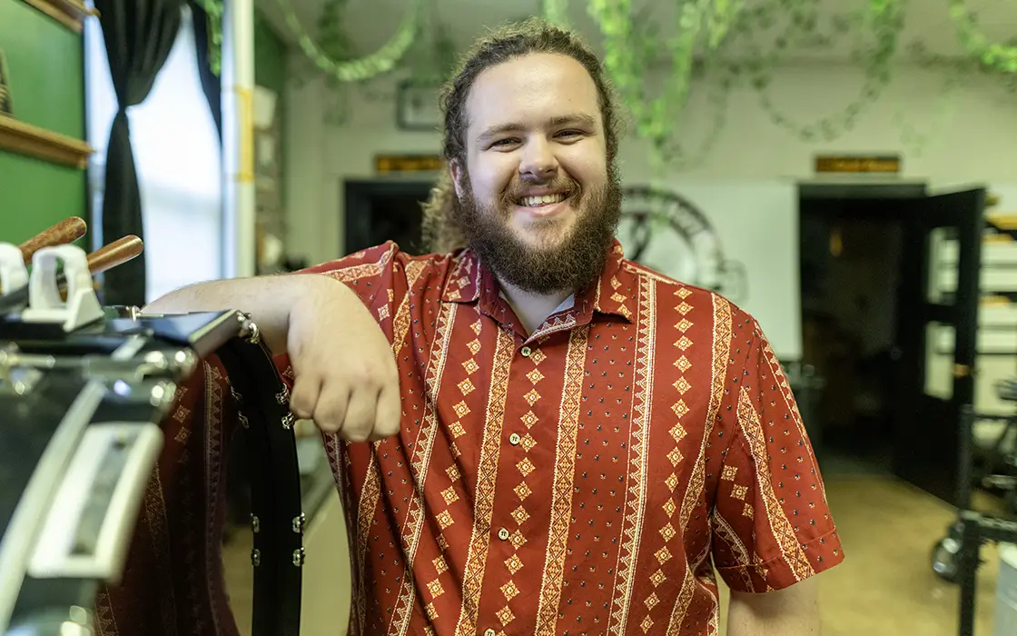 A man in a striped shirt smiles at the camera. His elbow is propped up on a drum.