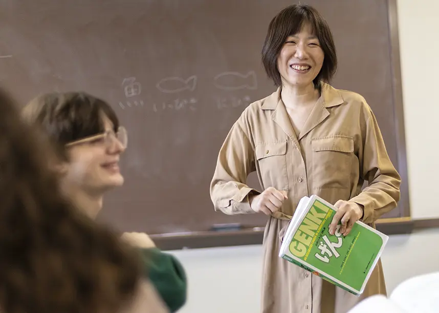 A Knox faculty members smiles and holds a. Japanese text book while standing in front of a chalkboard. A smiling student looking across the classroom is in the foreground.