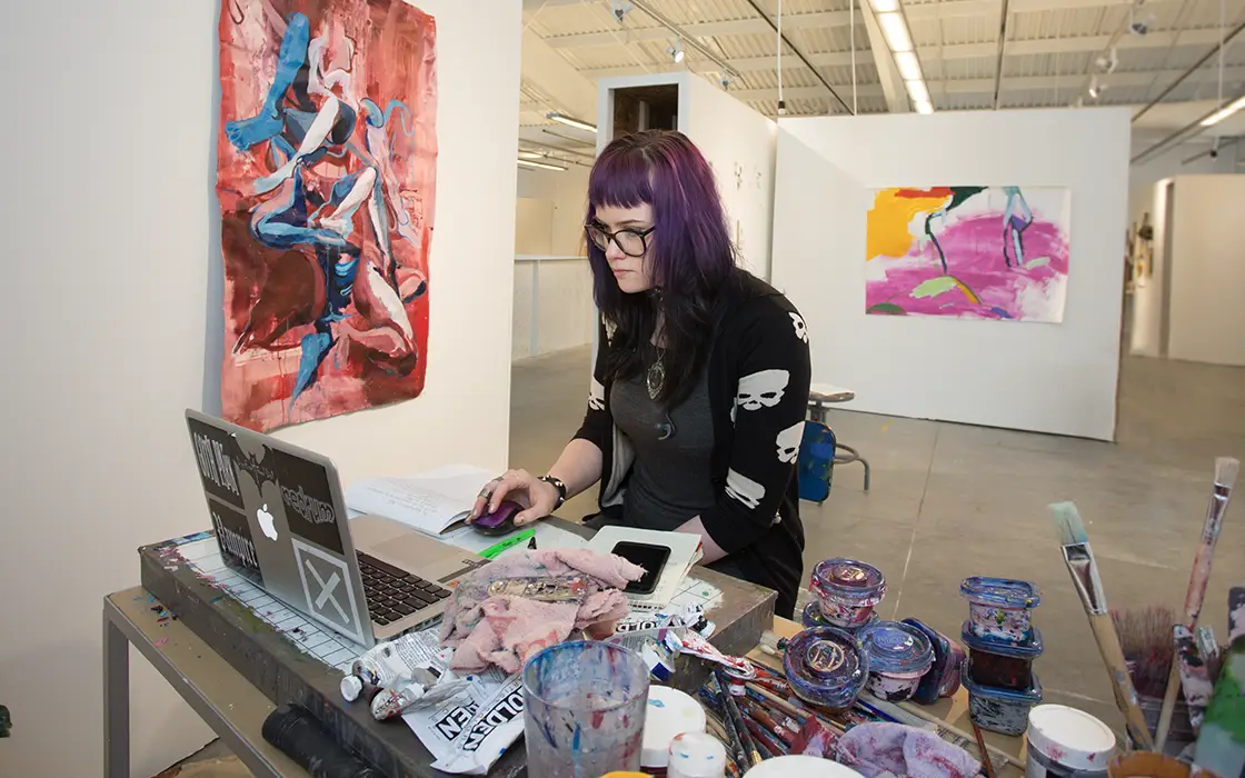 A student works on a laptop in a studio environment. She's surrounded by paintings, paints, and a sculpture.