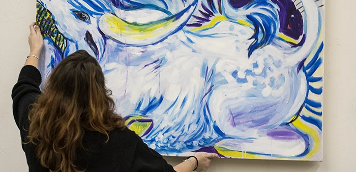 A person with long hair is photographed from behind as they hang a painting of an animal with blue and yellow undertones on the wall.