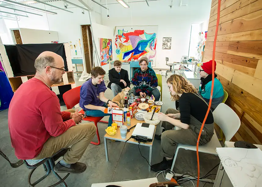 A group of six individuals and one dog gather around a table in a studio atmosphere. Snacks and drinks are spread out on the table.