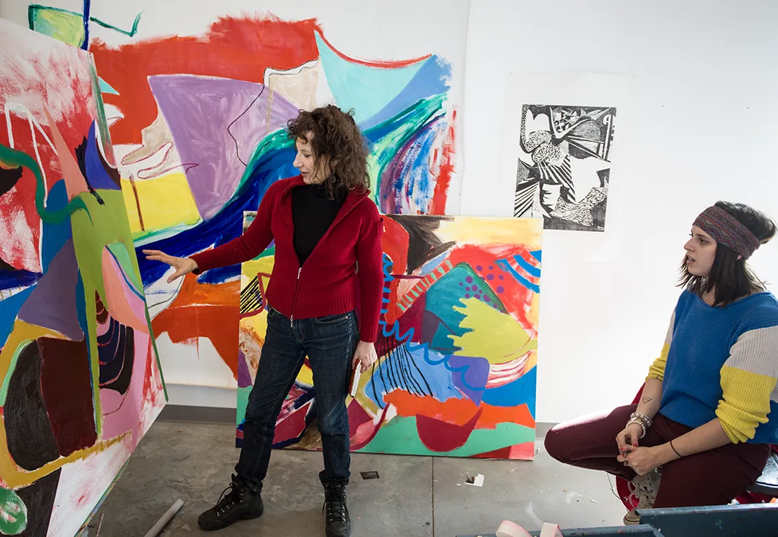 A professor looks at a painting and gestures toward it as she speaks to a student, who listens intently.