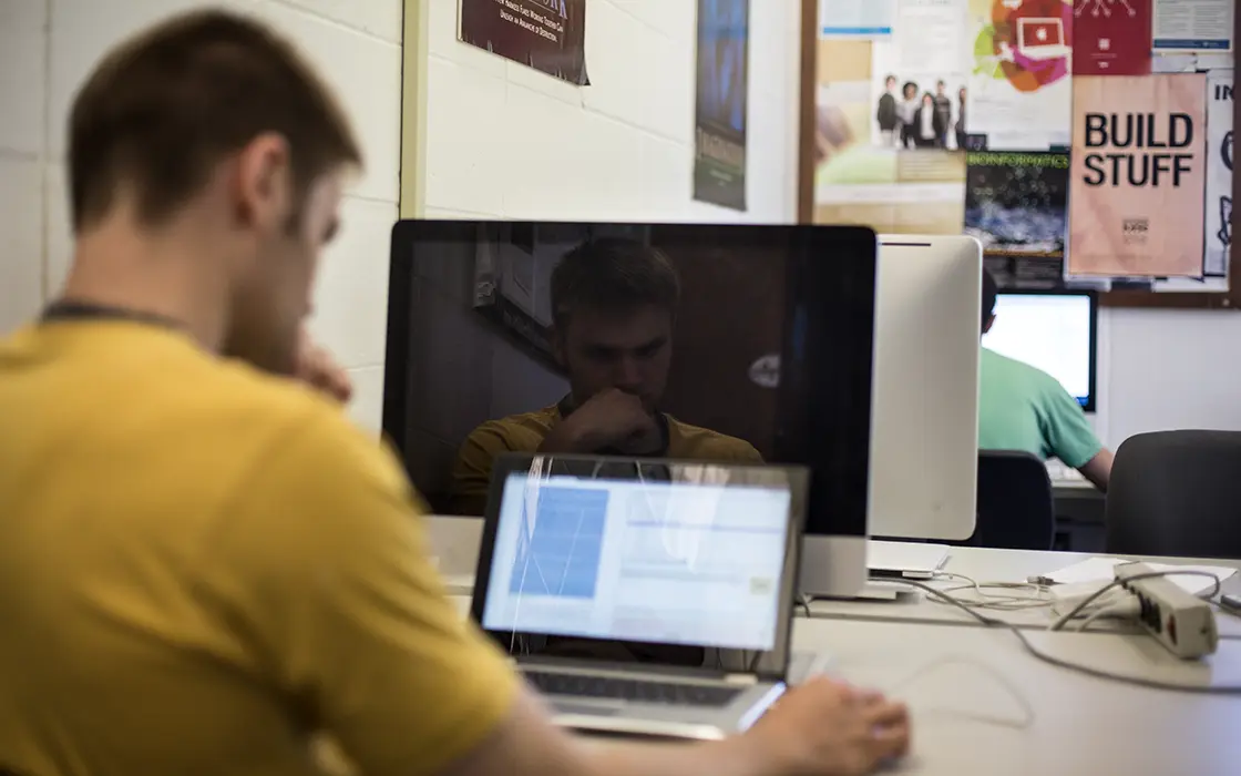 A student sits at a desk with two computers in front of him.