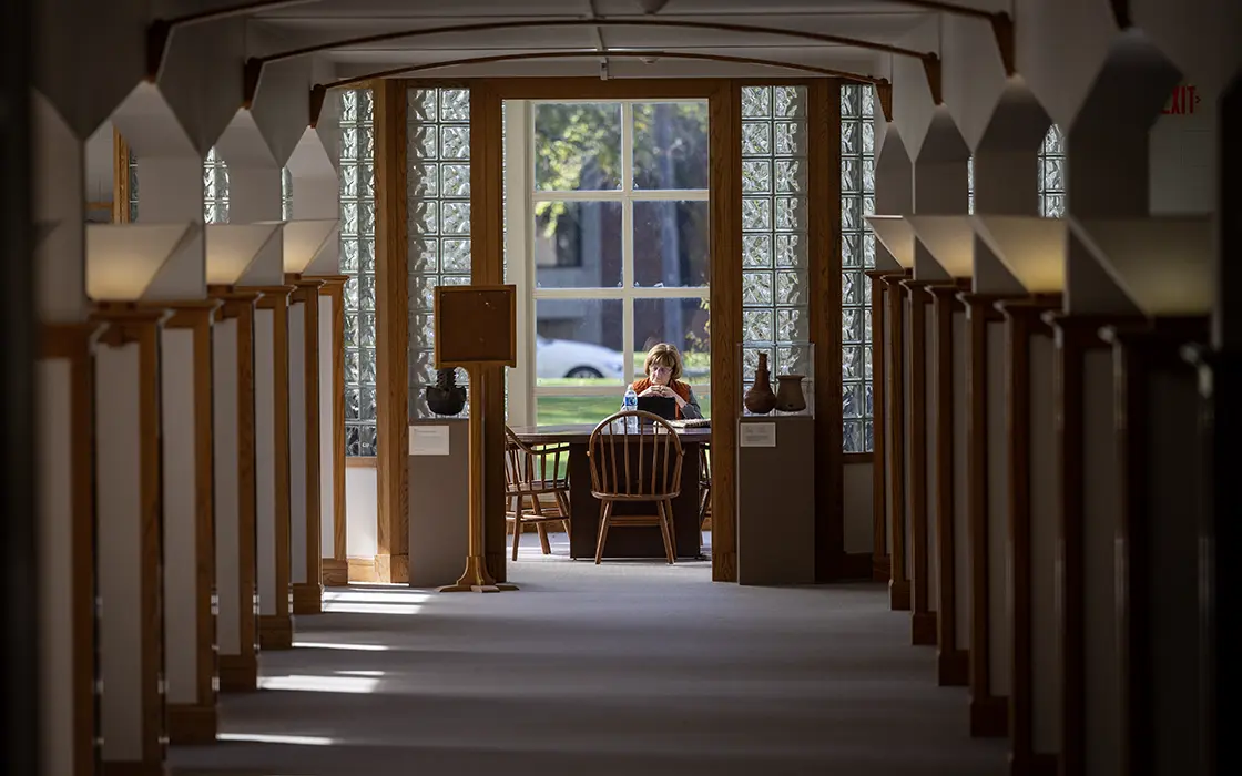 A person sits at a desk in the library, in front of windows. 