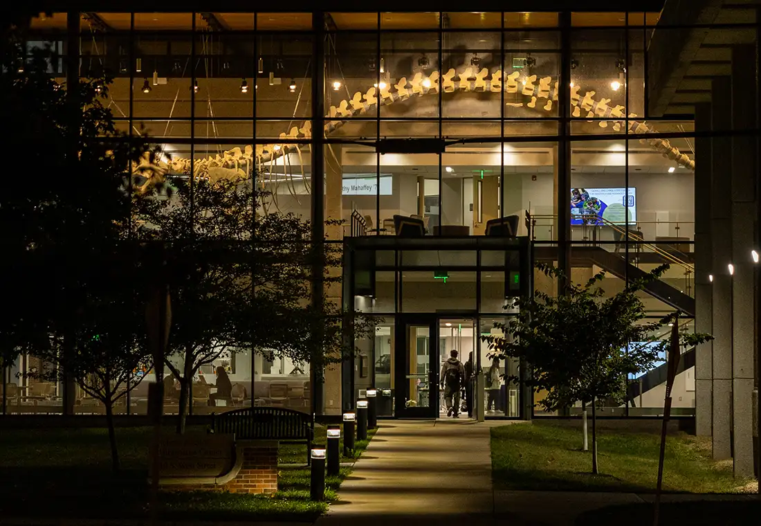Looking into a lit up building at night. A whale skeleton hangs from the ceiling of the second floor. Students are walking into the building