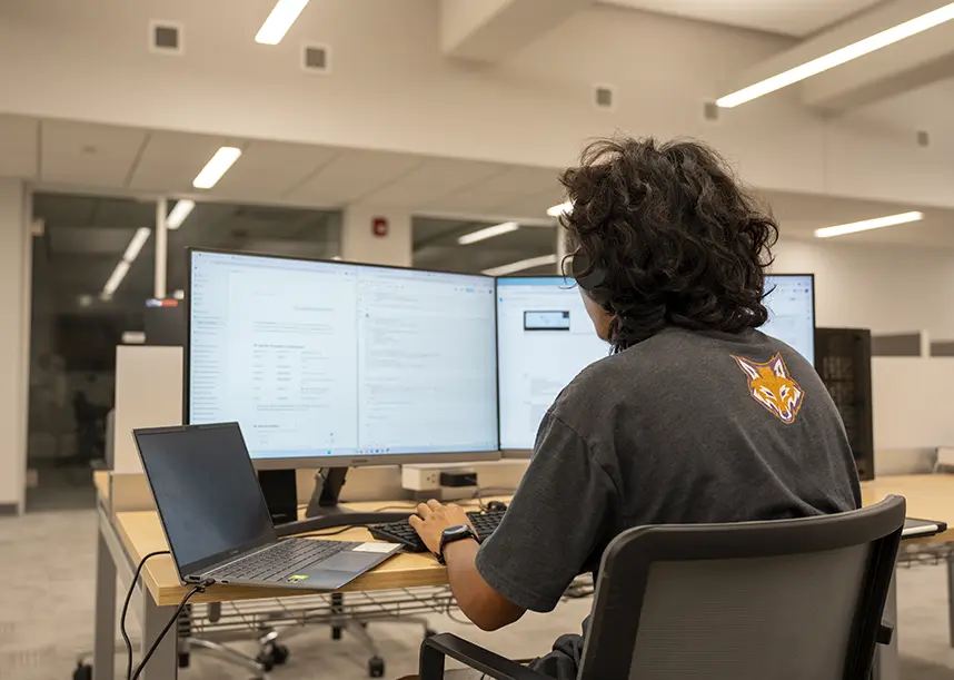 A student sits at two monitors working on a project.