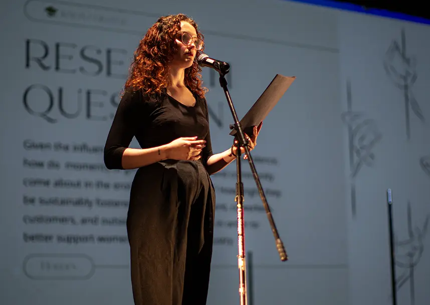 A student stands at the front of a stage, speaking into a microphone, with a large screen behind her.