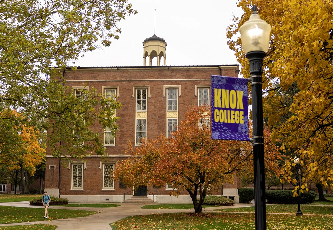 Old Main is pictured in early fall. A lightpost is in the foreground with a banner that reads Knox College.