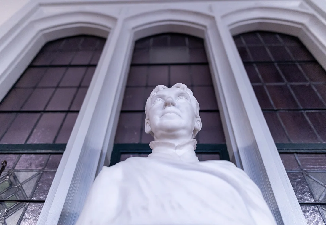 A white marble bust, photographed from the bottom looking up, sits in front of stained glass windows.