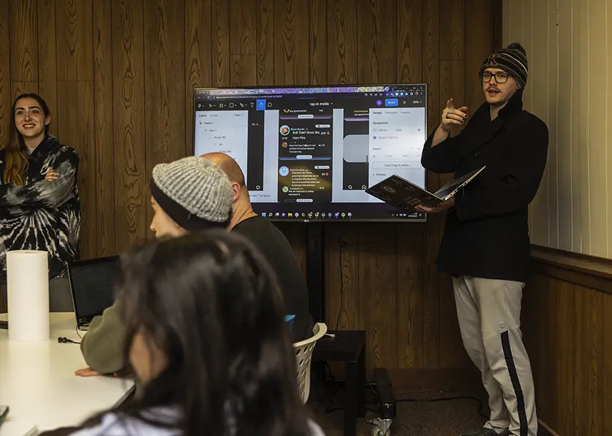 Two students stand in front of a classroom presenting on a screen between the two of them.