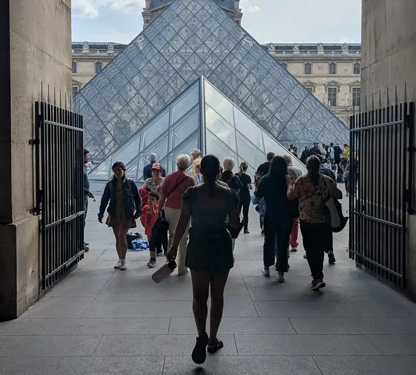 The Louvre Pyramid and the Louvre are visible beyond a gate. A small crowd of people is passing through.