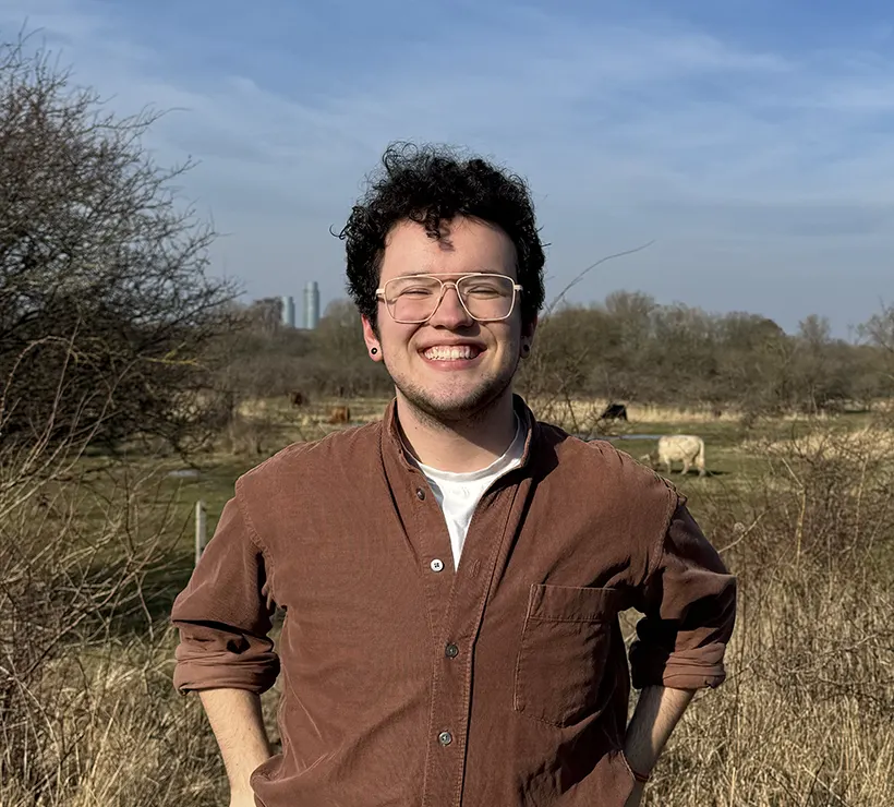 A student standing outdoors in an open space flashes a big smile at the camera; cows are visible over his shoulder.
