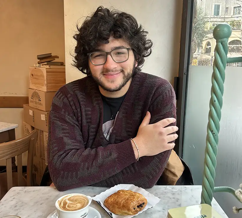 A student sits at a cafe table, next to a large window. In front of him on the table are a pastry and a latte.