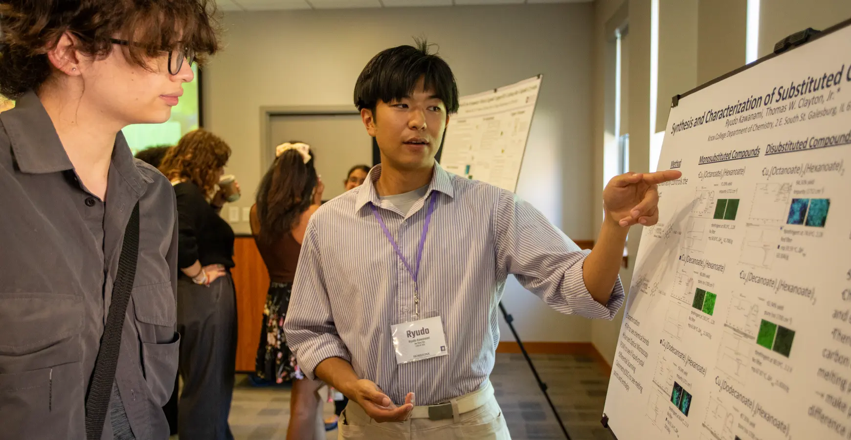 A student presents research to another student while standing in front of a poster.