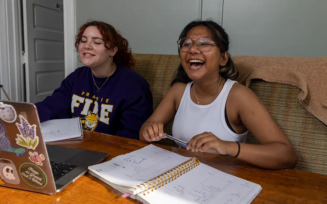 Two smiling students study together at a table. One is using a lapto, the other is looking at an open notebook.