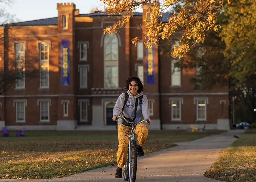 A smiling Knox student rides their back towards the camera on a fall afternoon. Old Main is in the background.