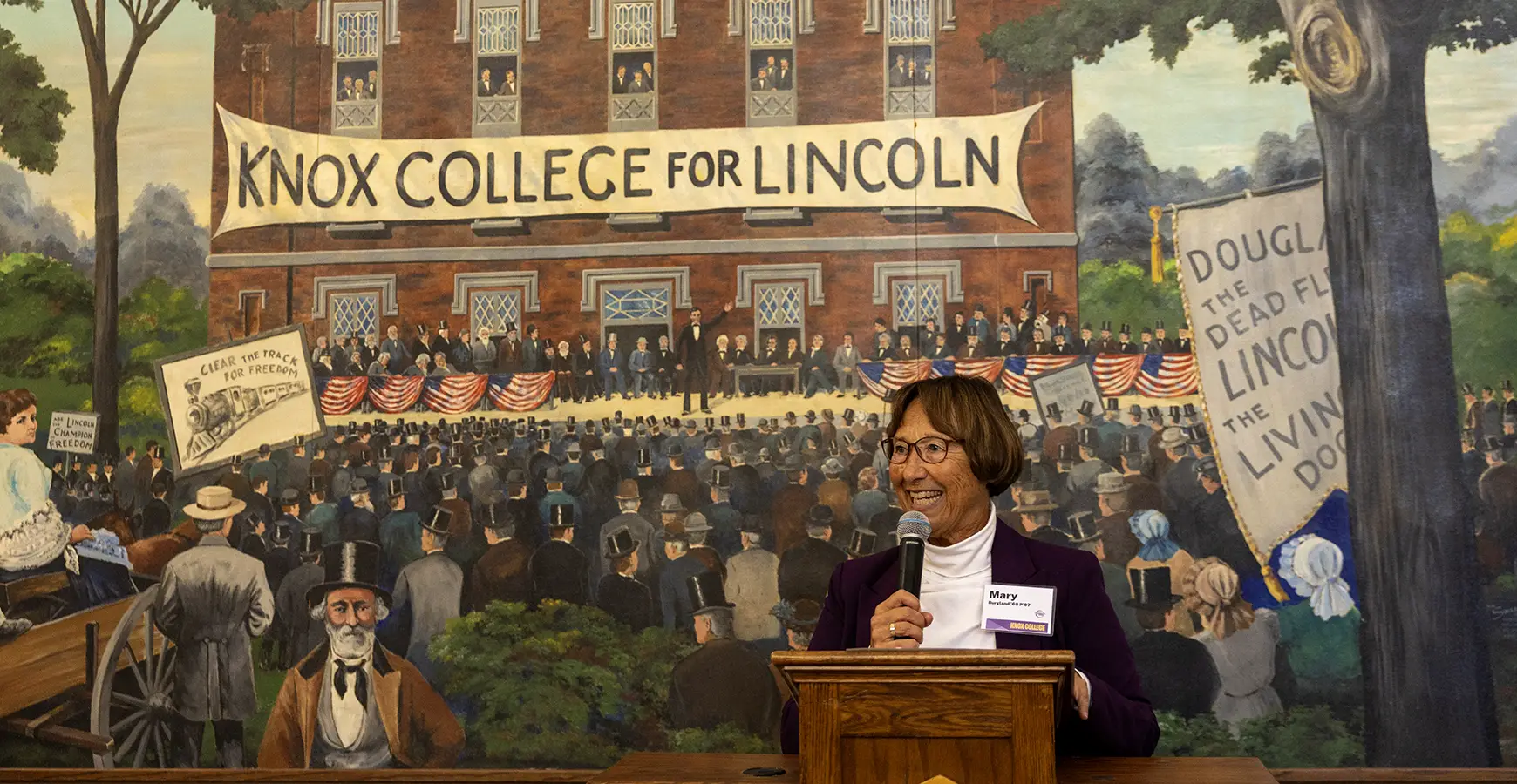 The president of the Fifty Year Club talks at a podium in front of a mural depicting the 1858 Lincoln-Douglas Debate.