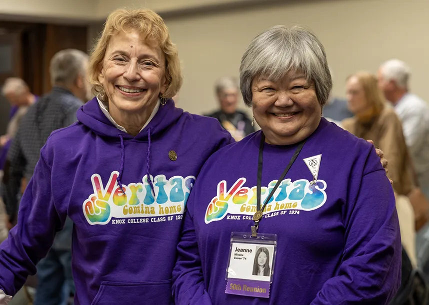 Two members of the Class of 1974 celebrate their 50th Reunion. They both are smiling at the camera and wearing purple sweatshirts that say, "Veritas, Coming Home" in tie-dye letters. 
