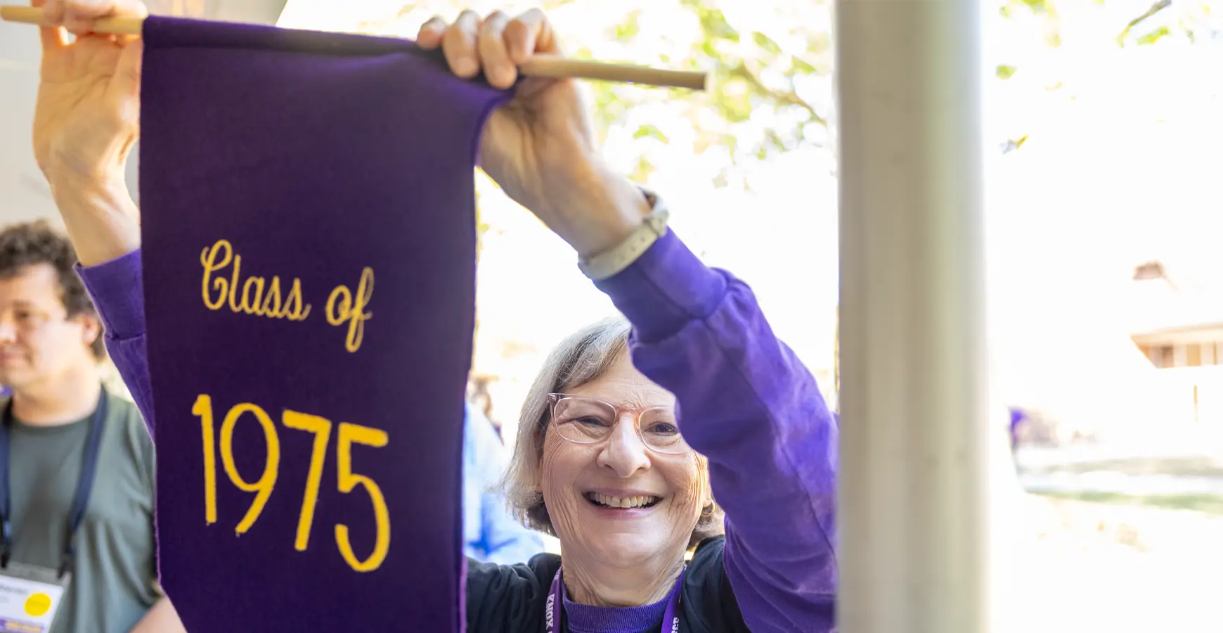 A woman smiles at the camera, holding up a purple banner that reads Class of 1975. 