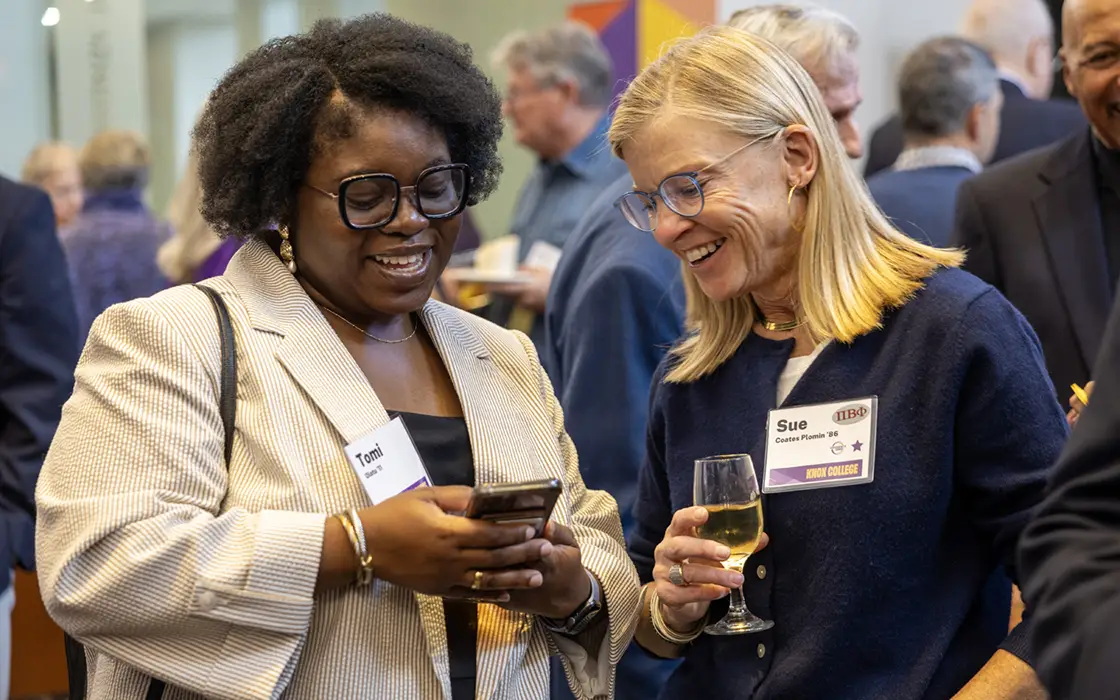 Two Knox alumni talk at a reception. One is showing their phone to the other, who holds a glass of wine.