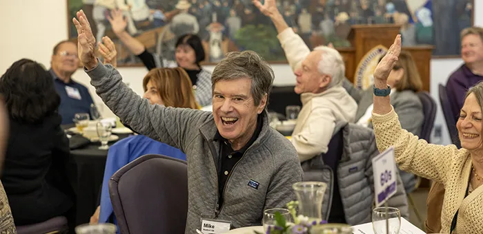 Smiling alumni sitting at circular tables smile and raise their hands during a reception.