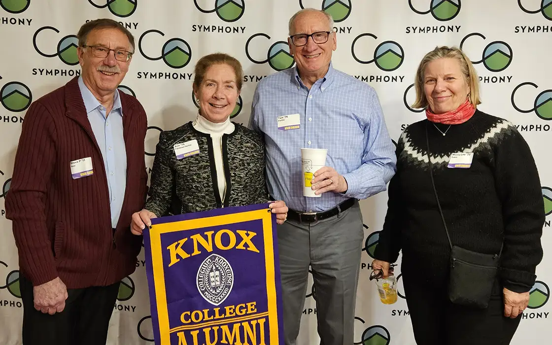 Four members of the Colorado Knox Club stand in front of a Colorado Symphony. One holds a Knox College alumni flag.