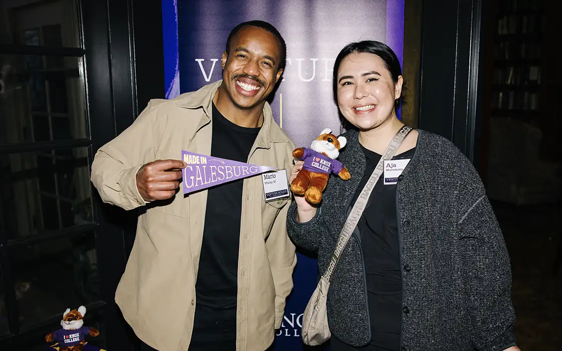 Two Knox alumni smile at the camera with a Galesburg pennant and Knox Fox plushie in front of a backdrop that says "Venture Bodly."